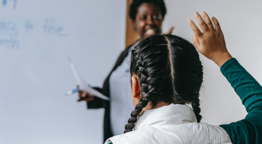 Teacher standing by a white board, and pupil with hand in the air.